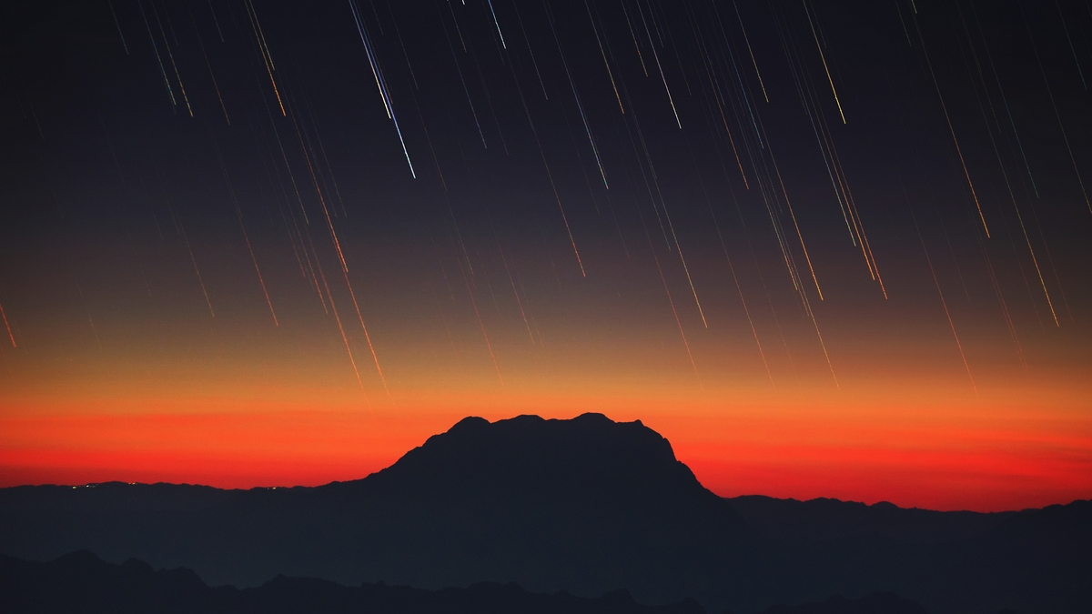 Star trails falling down over mountain range with sunset sky gradient.