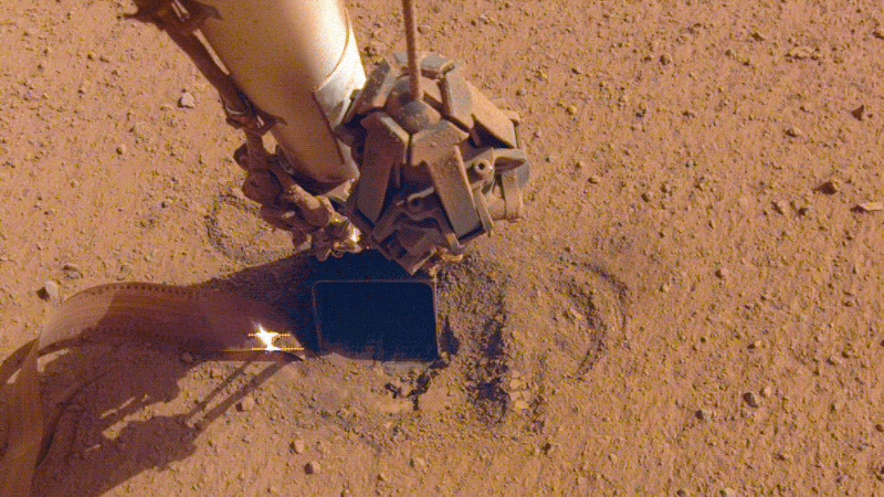 NASA's InSight lander retracting its robotic arm, revealing the spot where the mole is now completely buried.  (Gif: NASA/JPL-Caltech/Gizmodo)