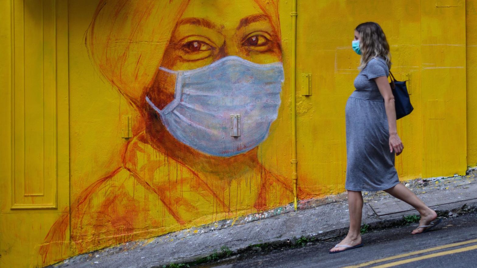 A pregnant woman wearing a face mask walks past a street mural in Hong Kong, on March 23, 2020, (Photo: Anthony Wallace/AFP, Getty Images)