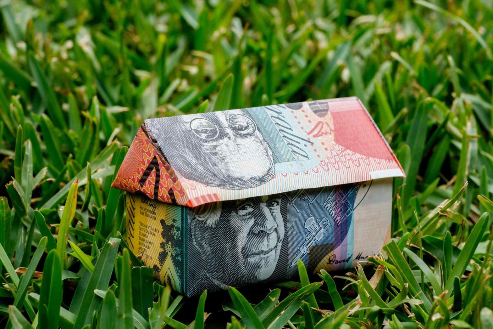 Folded Australian dollar notes make up a tiny house on a green lawn