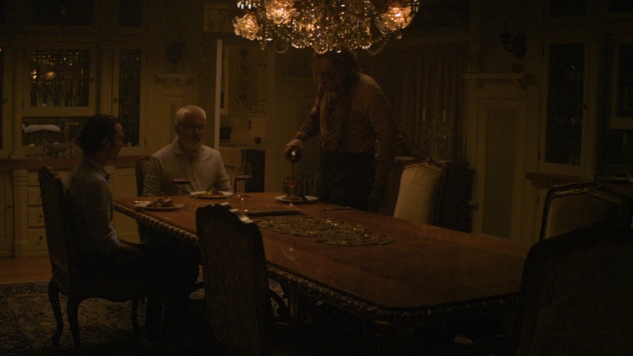 Nic Cage pours a glass of wine for himself, with a man and a younger man sitting at the table waiting for him