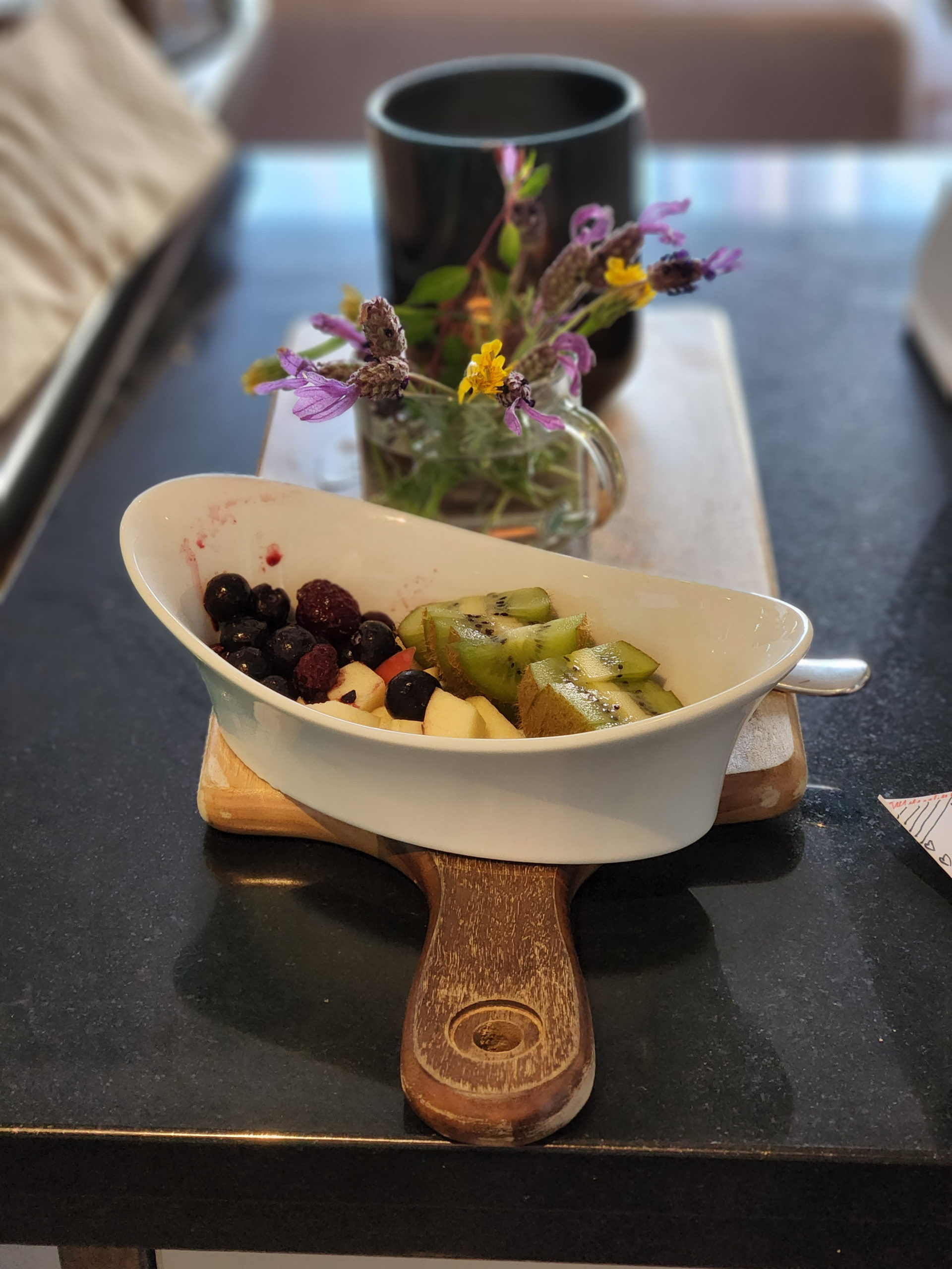 A bowl of sliced fruit on a wooden board in front of a vase and flowers and a candle.