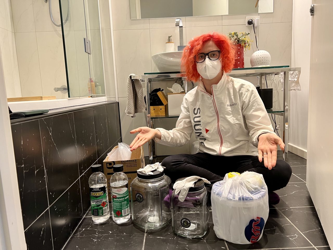Preserving a human organ: A person with orange hair in old clothes gesturing at a white bucket, two empty jars and some methylated spirits bottles. 