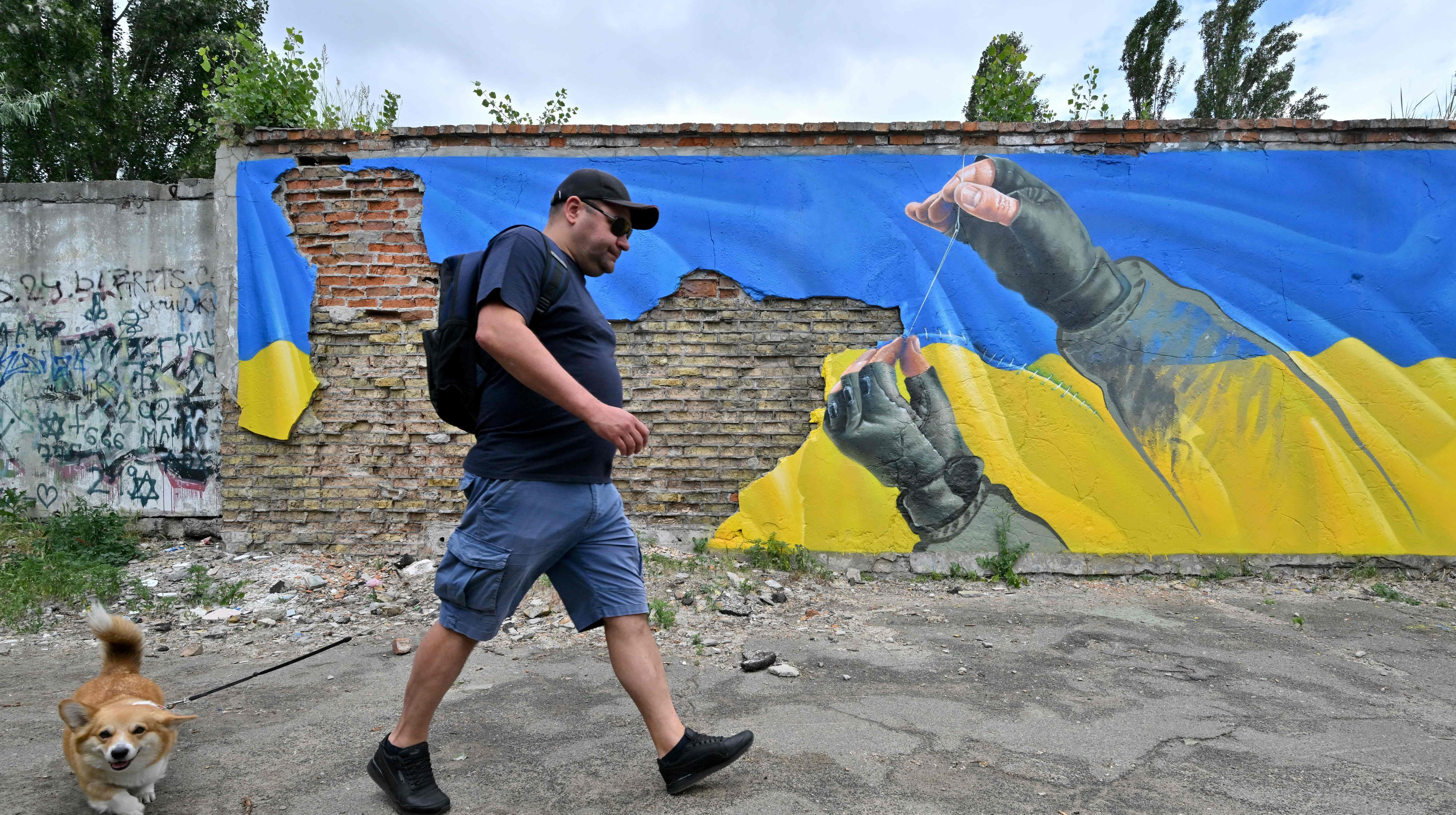 A man walks with his dog past a mural by street artist Sasha Korban depicting hands of a military man sewing together parts of the Ukrainian flag, in Kyiv on June 14, 2022. (Photo: SERGEI SUPINSKY/AFP, Getty Images)