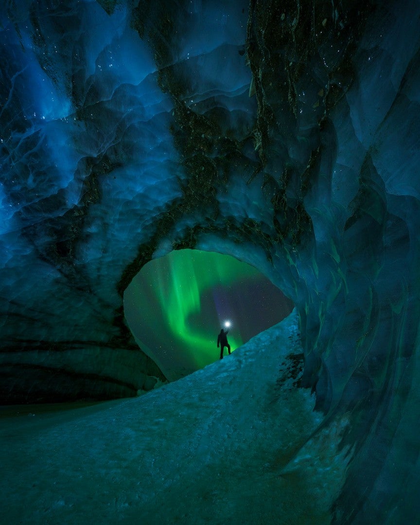 A person silhouetted under the Northern Lights, looking out from an ice cave. (Photo: David Erichsen)